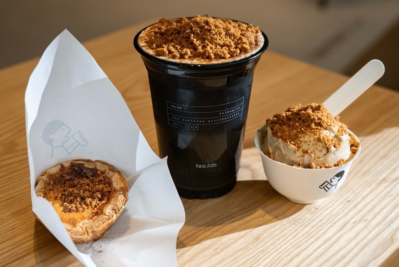 Heytea's three UK exclusives on a wooden table: a croissant egg tart topped with biscuit crumbs, a tall Tea Lab cup crowned with biscuit crumble and a cup of milk tea ice cream with more crumbs.