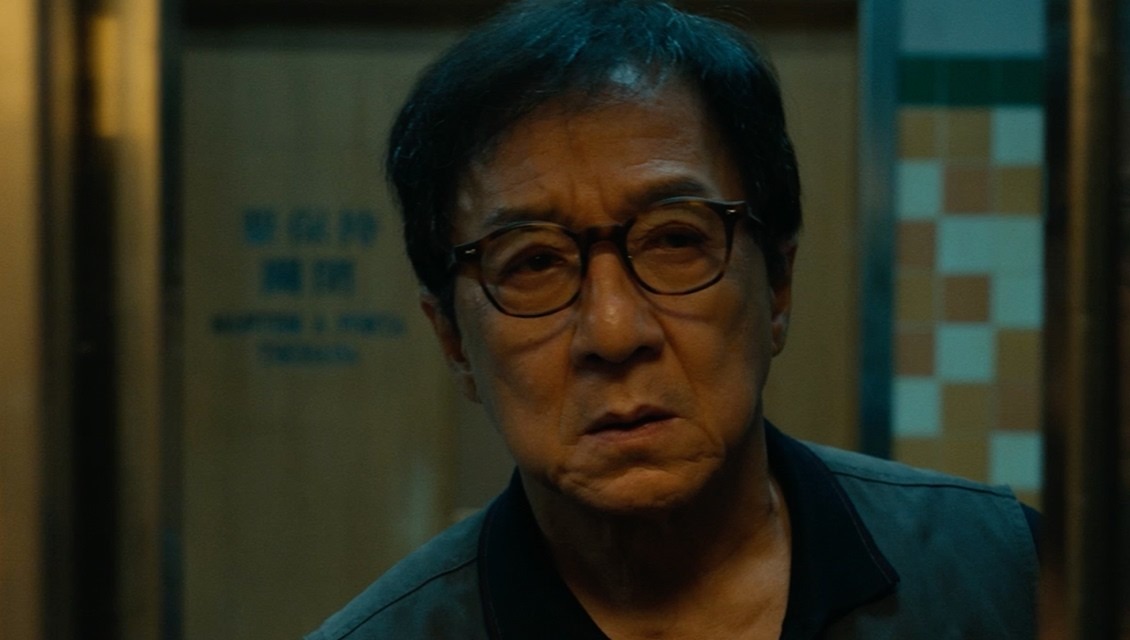Jackie Chan in glasses and a dark vest, frowning in a dim corridor with tiled walls, in a still from The Shadow's Edge trailer.