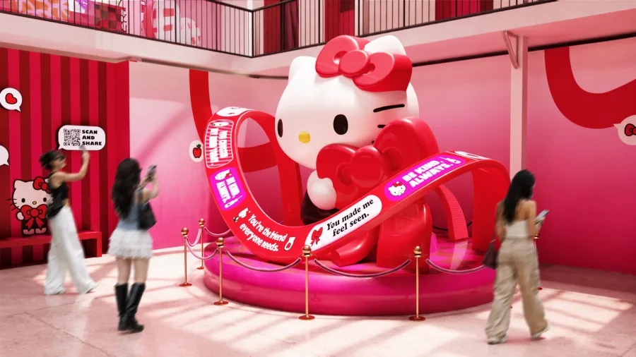 Render of the exhibition: a giant white Hello Kitty figure with a red bow sits on a pink platform wrapped in red ribbons printed with messages, as visitors take photos in a pink and red room.