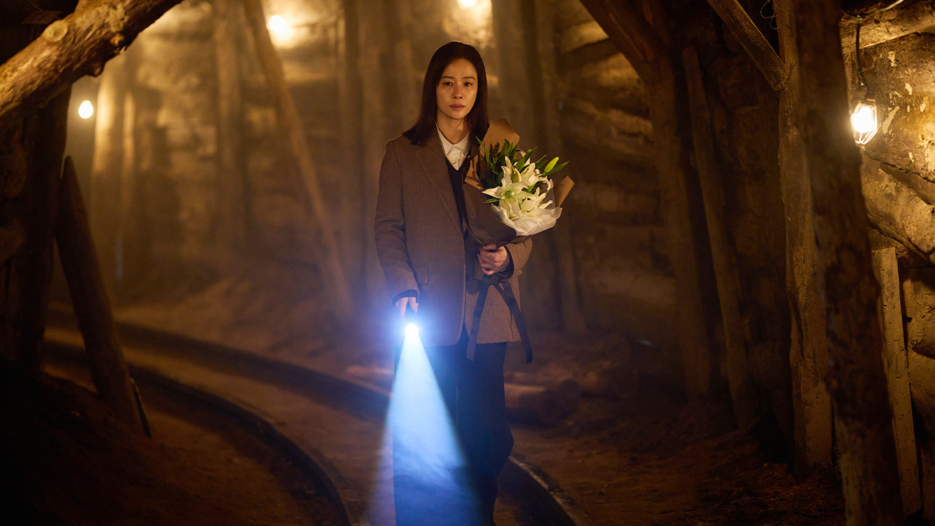 Film still: a woman in a long coat walks through a timber-propped mine tunnel holding white lilies and a torch whose blue beam lights the ground ahead.