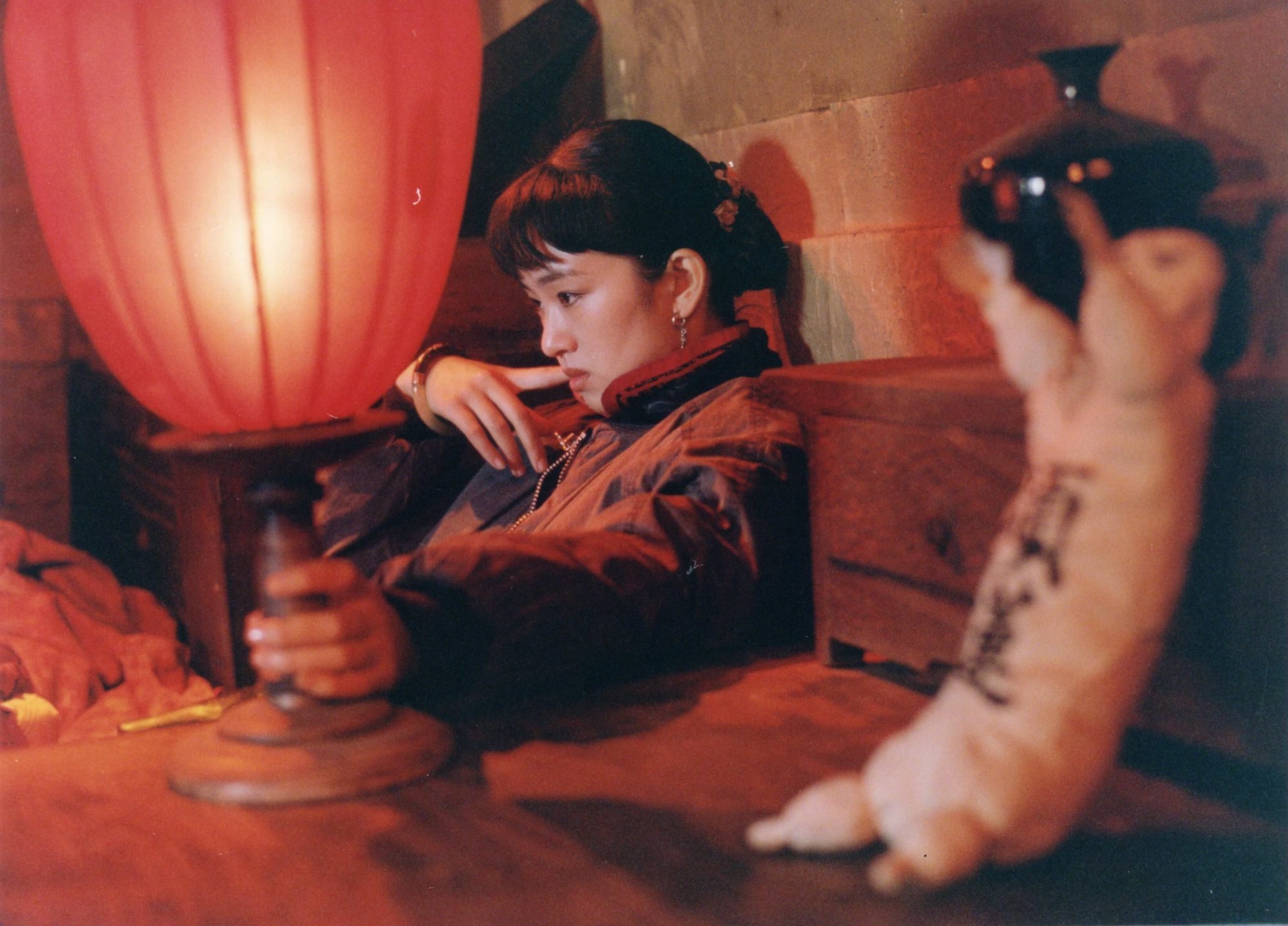 Film still: a woman in a dark padded jacket leans back against a wooden chest, one hand on the stand of a large glowing red lantern, in a dim room lit red.