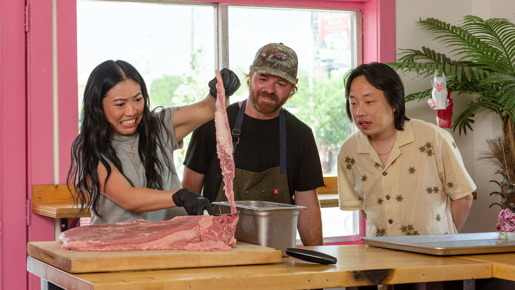 Awkwafina holds up a long strip of raw beef over a butcher's block while two men watch, in a kitchen with pink window frames
